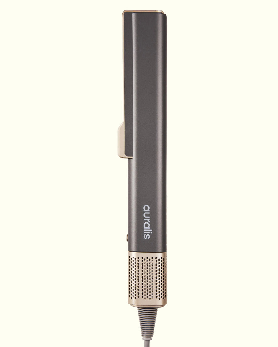 Alisador de aire Aeroliss™ de Auralis en gris grafito con detalles rosa dorado/champán, de pie sobre fondo crema, vista de perfil cerrada: cuerpo rectangular alargado con un clip curvo en la parte superior, el logo "auralis" grabado verticalmente en el mango, un pequeño botón lateral, y base inferior con malla metálica rosa dorado y el cable de alimentación.