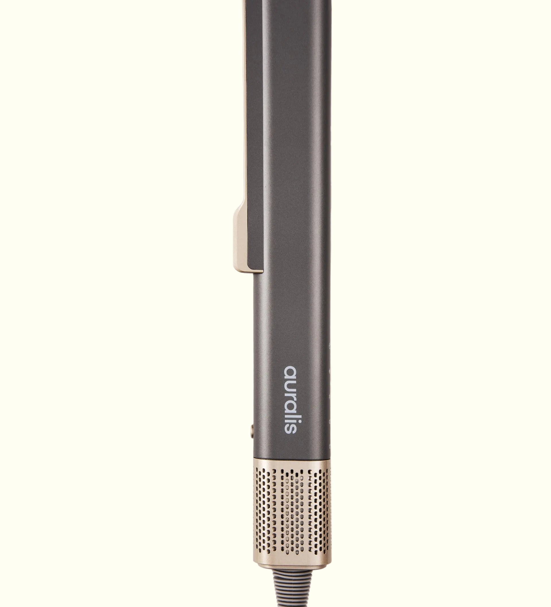 champán, de pie sobre fondo crema, vista de perfil cerrada: cuerpo rectangular alargado con un clip curvo en la parte superior, el logo "auralis" grabado verticalmente en el mango, un pequeño botón lateral, y base inferior con malla metálica rosa dorado y el cable de alimentación.