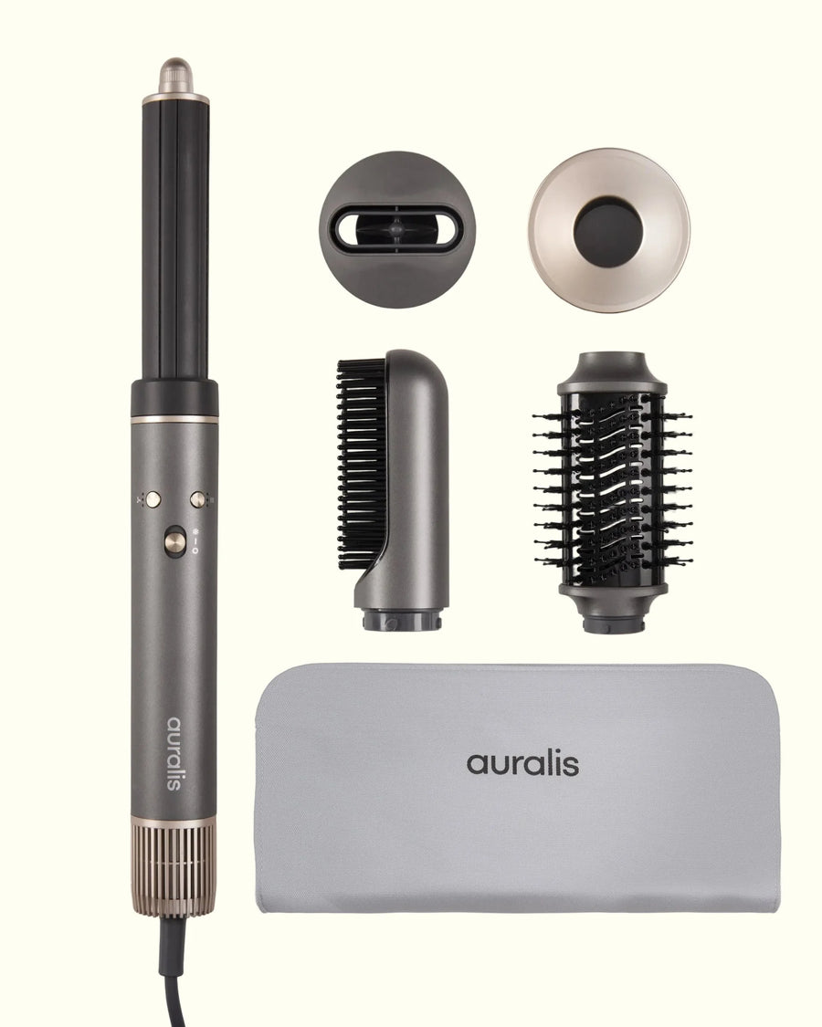 Set completo del secador multiestilo Aeroflow™ de Auralis en color gris grafito, fotografiado sobre fondo crema: el cuerpo principal con boquilla difusora redonda a la izquierda; arriba a la derecha dos tapas de accesorio, una ovalada negra y una circular en tono champán; en el centro un cepillo redondo térmico de cerdas negras y un accesorio concentrador de boquilla estrecha; abajo una funda de viaje de tela gris con el logo "auralis" bordado.