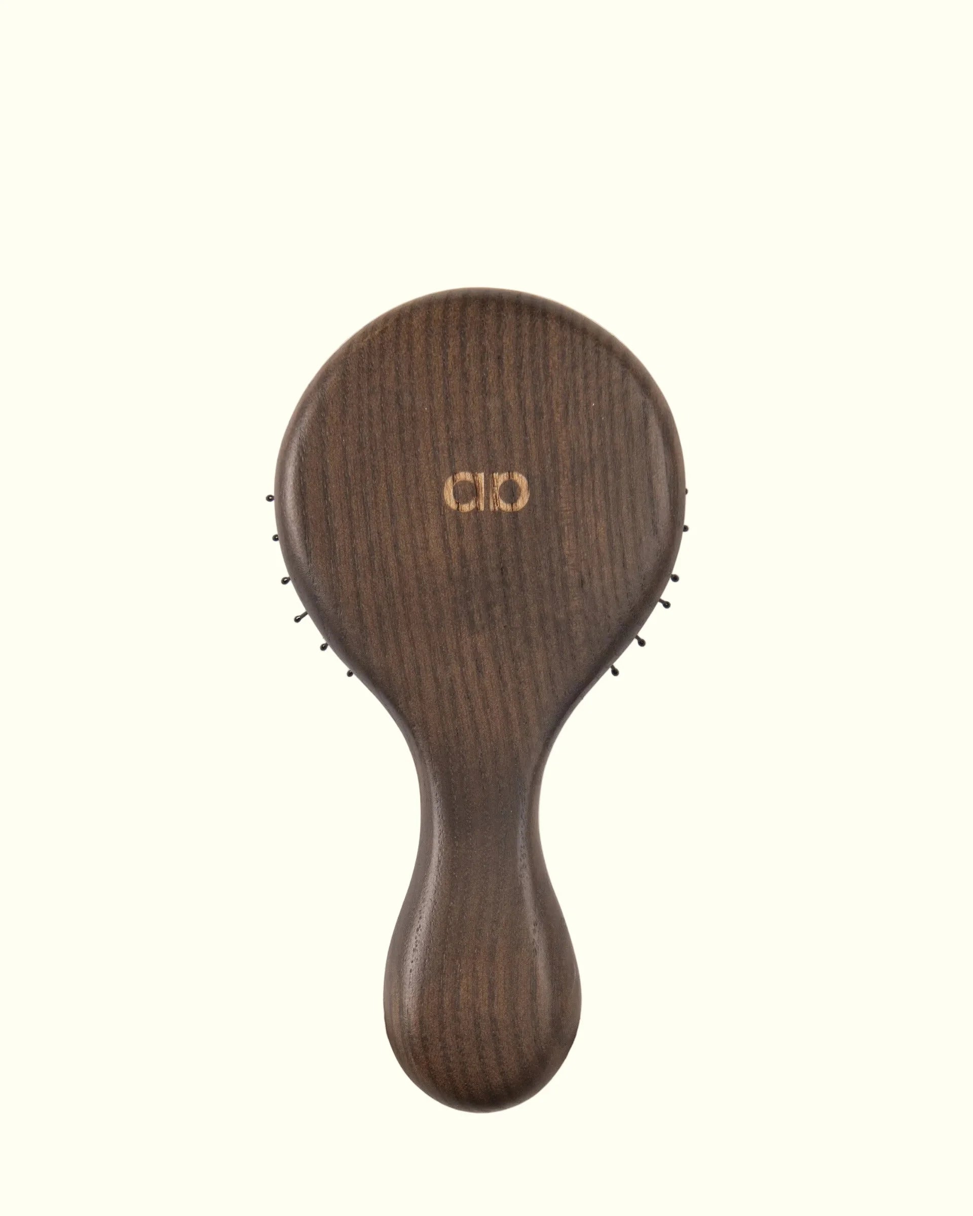 Cepillo técnico compacto Auralis Icon Travel™, vista trasera sobre fondo crema: dorso de madera oscura con veta natural visible y el logo de Auralis grabado en dorado en el centro del cabezal; se asoman las puntas de las cerdas negras alrededor del borde; mango redondeado de la misma madera.
