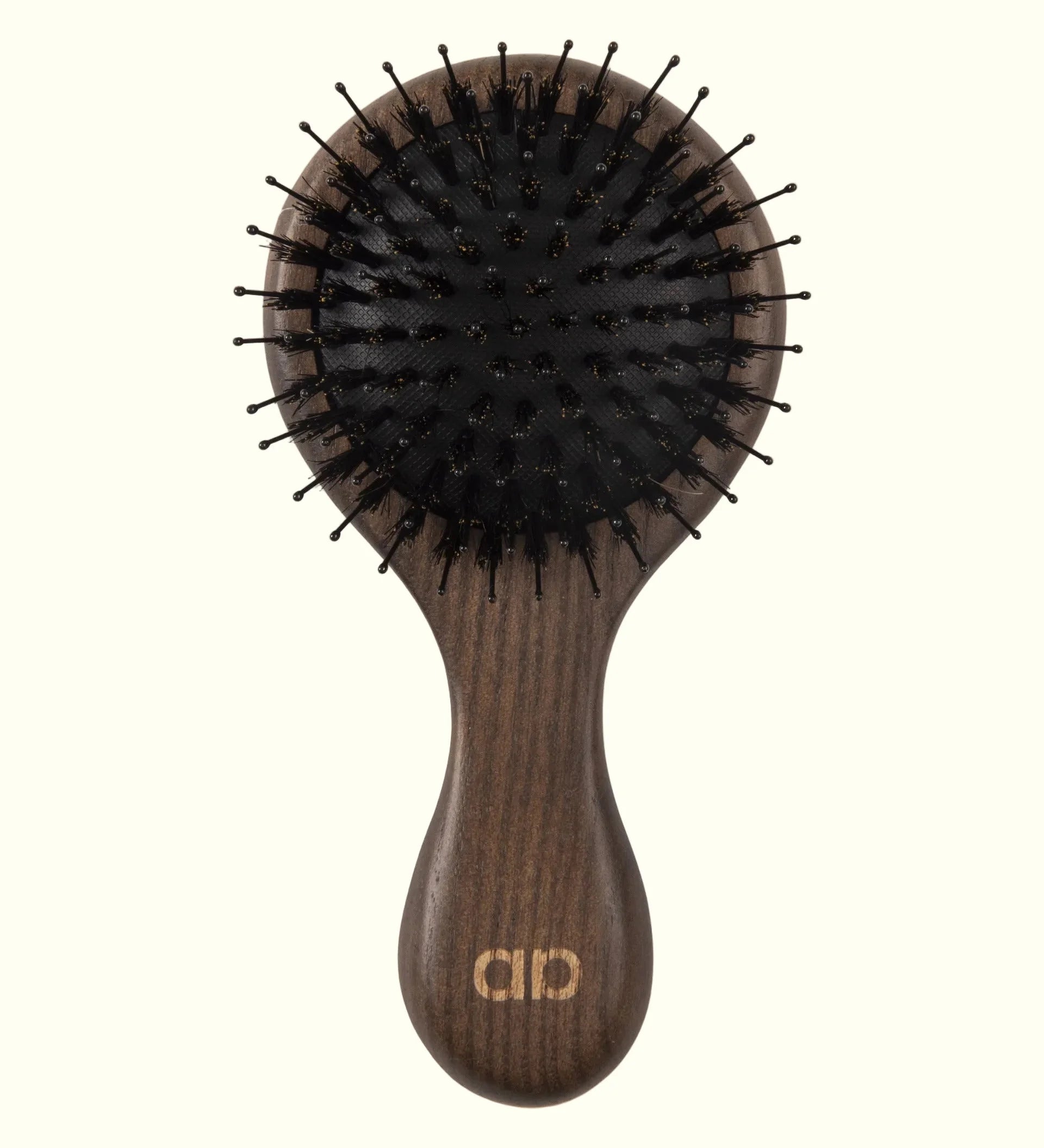 Cepillo técnico premium Auralis Icon™, vista frontal sobre fondo crema: cabezal ovalado grande con base de cojín flexible y cerdas negras densas tipo pin; mango largo y redondeado de madera oscura con veta visible, con el logo de Auralis grabado en dorado en la base del mango.