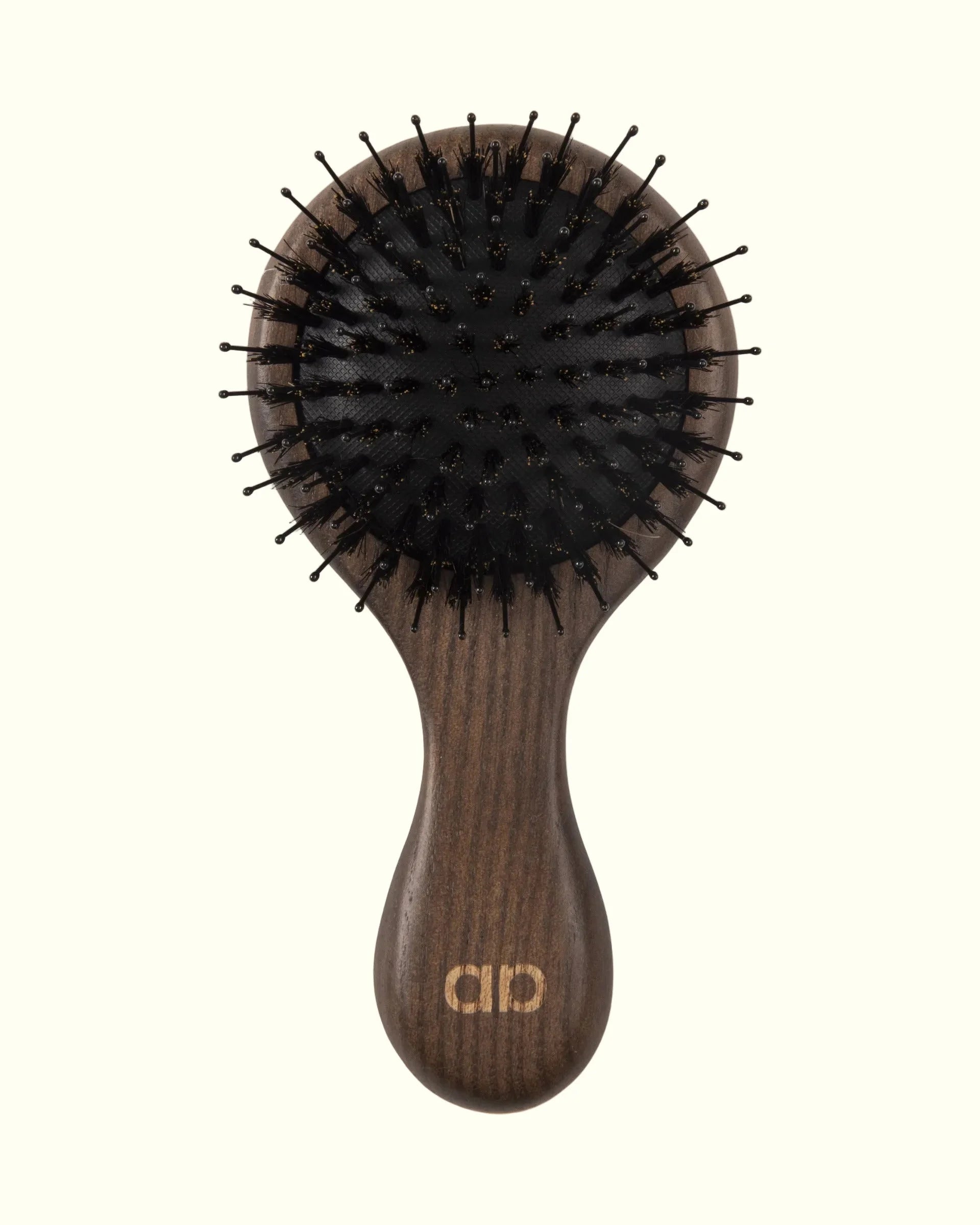 Cepillo técnico premium Auralis Icon™, vista frontal sobre fondo crema: cabezal ovalado grande con base de cojín flexible y cerdas negras densas tipo pin; mango largo y redondeado de madera oscura con veta visible, con el logo de Auralis grabado en dorado en la base del mango.