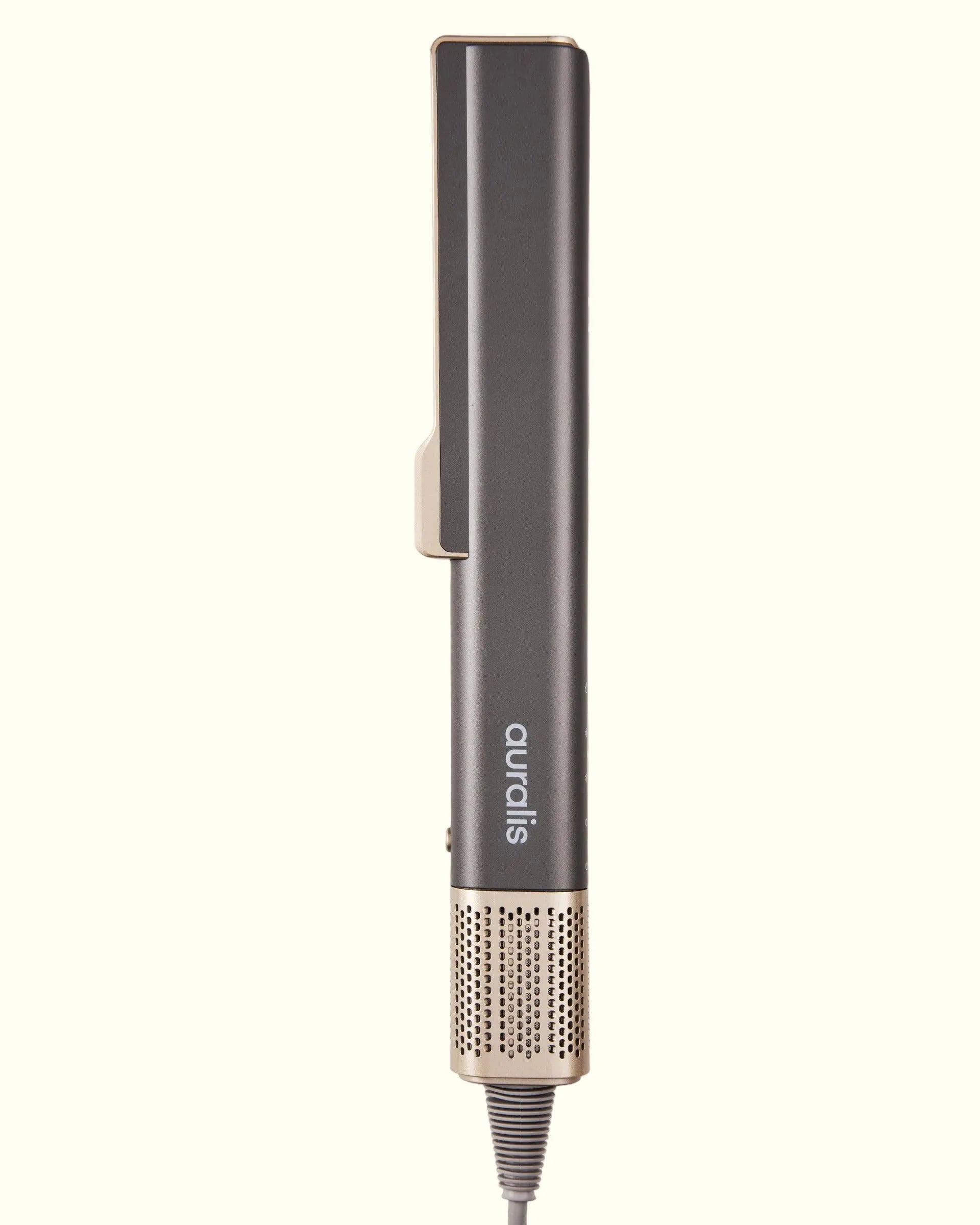 champán, de pie sobre fondo crema, vista de perfil cerrada: cuerpo rectangular alargado con un clip curvo en la parte superior, el logo "auralis" grabado verticalmente en el mango, un pequeño botón lateral, y base inferior con malla metálica rosa dorado y el cable de alimentación.
