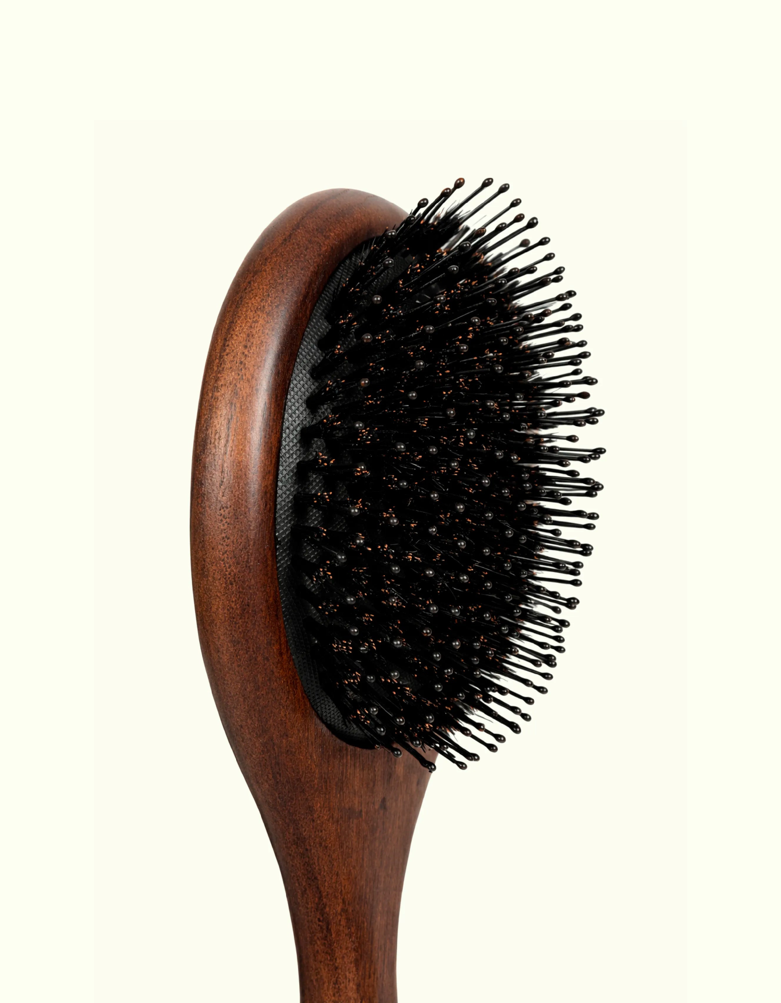 Fotografía de primer plano del cabezal del cepillo técnico Auralis Icon™: base de cojín acolchado con textura de malla y cerdas negras tipo pin con puntas redondeadas color cobre, montadas en un cuerpo de madera de tono caoba rojizo, sobre fondo crema claro.