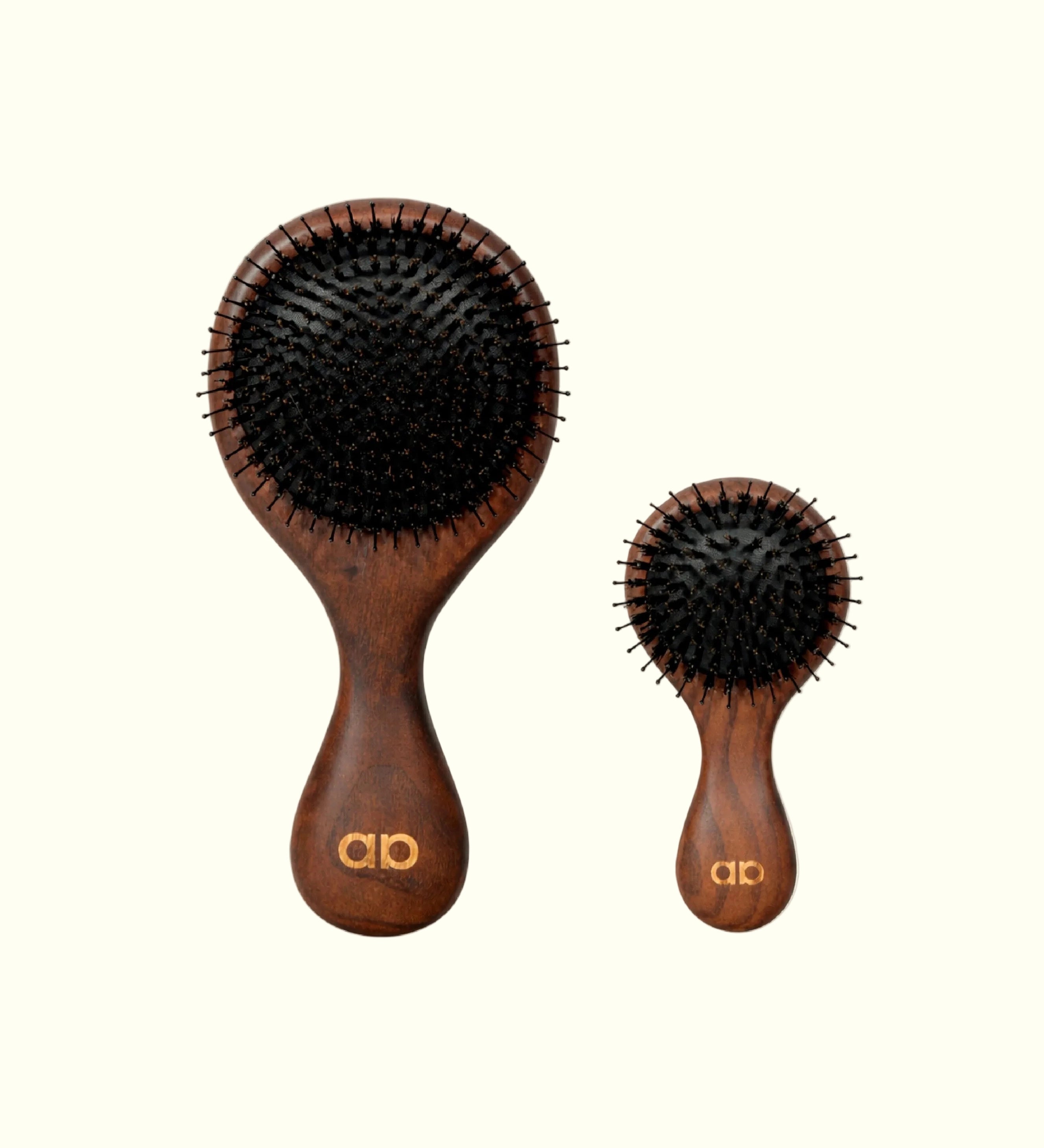Comparación de tamaño entre los dos cepillos técnicos Auralis: a la izquierda el modelo Icon de tamaño completo, con cabezal ovalado grande de cerdas negras y mango de madera oscura con el logo dorado "ab"; a la derecha, más pequeño, el modelo Icon Travel con el mismo diseño en versión compacta, ambos sobre fondo crema.