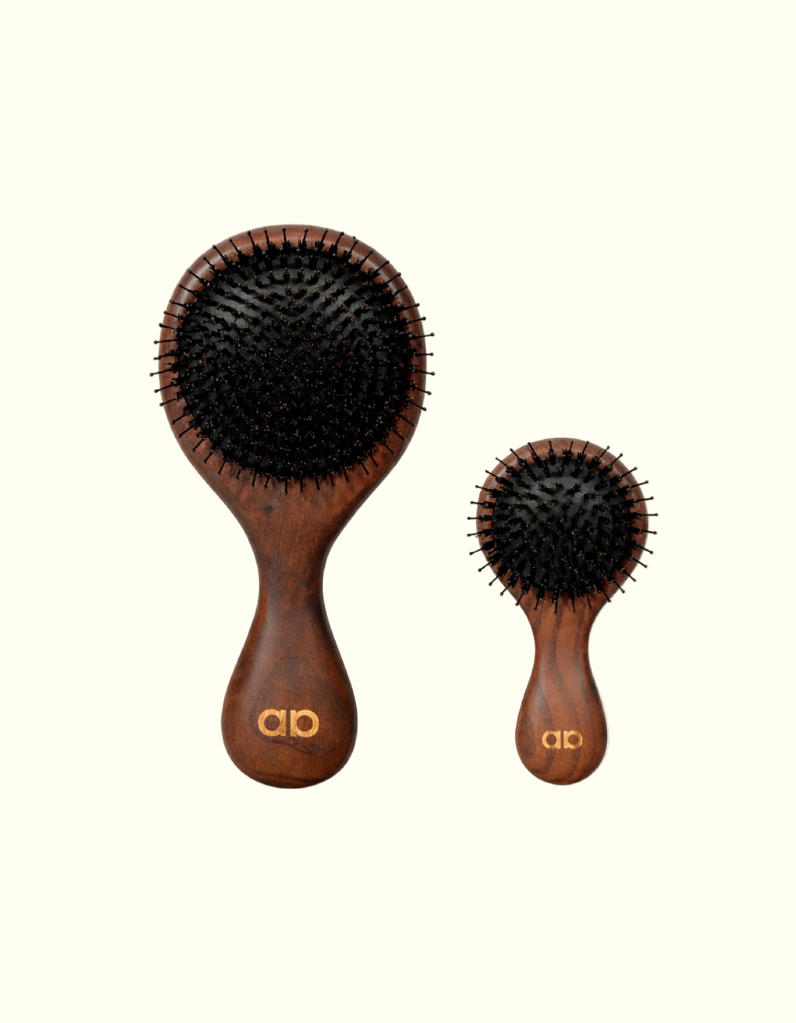 Comparación de tamaño entre los dos cepillos técnicos Auralis: a la izquierda el modelo Icon de tamaño completo, con cabezal ovalado grande de cerdas negras y mango de madera oscura con el logo dorado "ab"; a la derecha, más pequeño, el modelo Icon Travel con el mismo diseño en versión compacta, ambos sobre fondo crema.